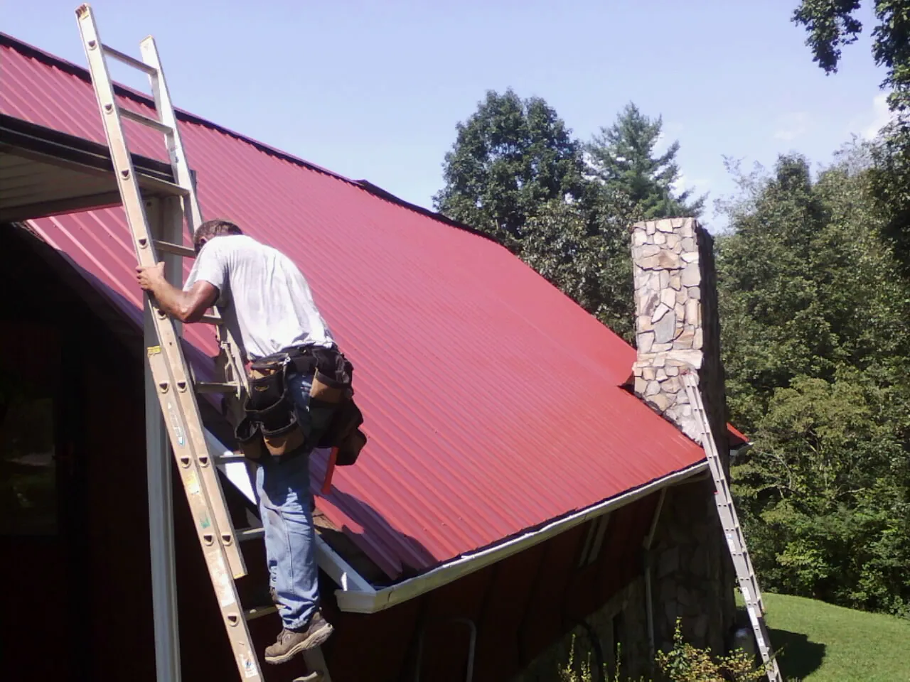 Roofer on a ladder beside a red metal roof and stone chimney — Soffit & Fascia Repair by Roofing Brothers in Rimrock